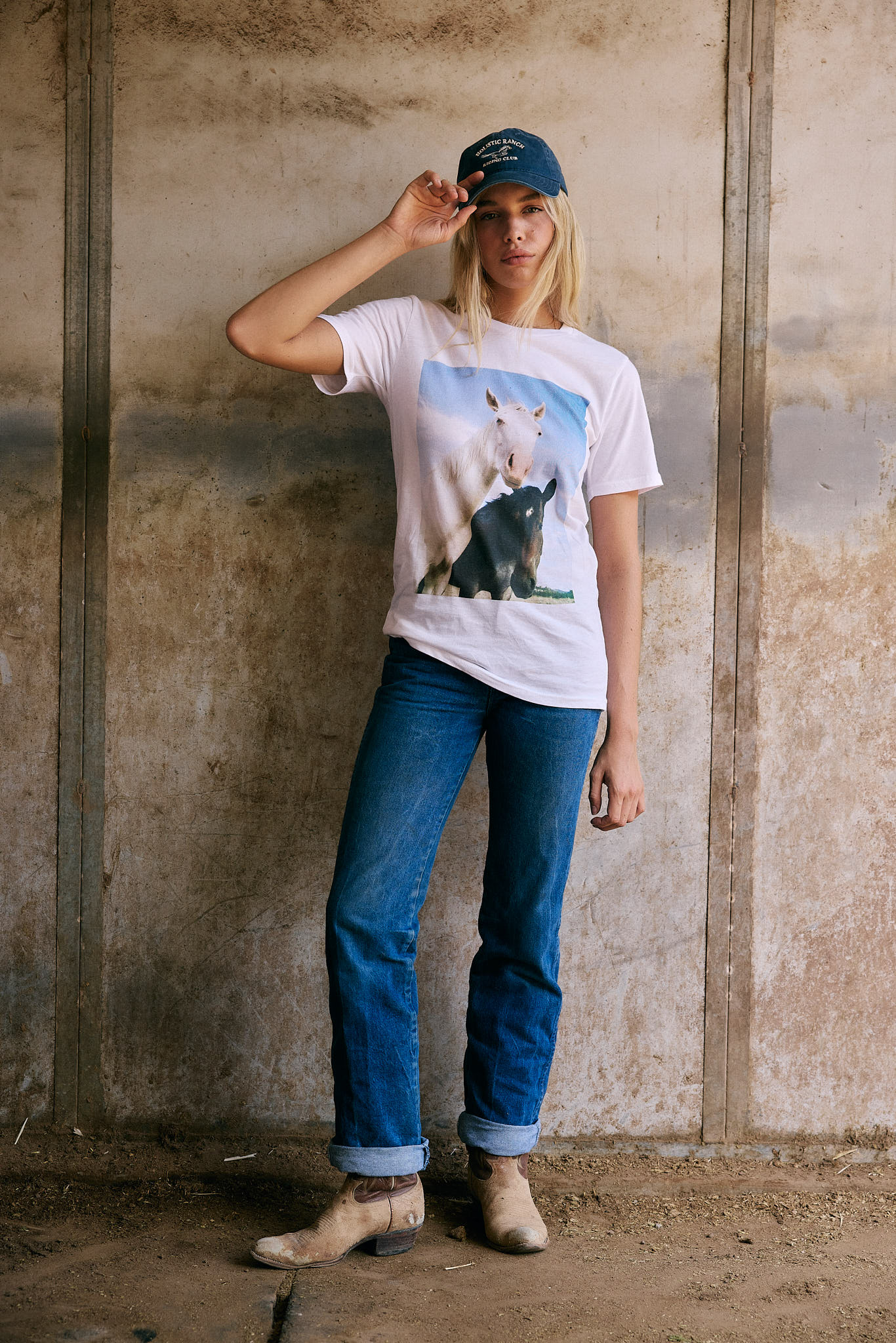 Model wearing Holistic Ranch Giveback Tee with horse graphic, photographed outdoors by a rocky creek. Vintage ranchwear T-shirt supporting horses in need.