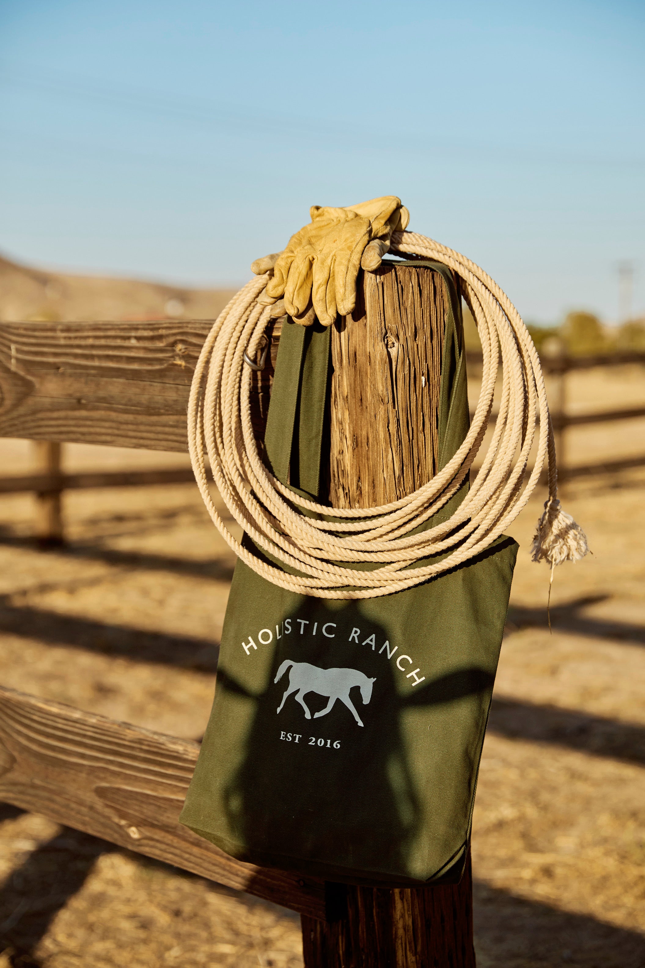 A Holistic Ranch Tote crafted from green organic certified cotton, featuring a horse logo and "Holistic Ranch, EST 2016" text, hangs on a wooden fence. A coiled rope and a pair of gloves rest on top, with a sunny ranch landscape in the background.
